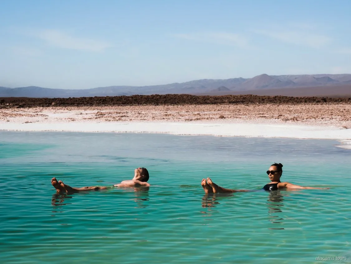 Passeios no Atacama, dois visitantes flutuando numa laguna salgada turquesa com montanhas ao fundo