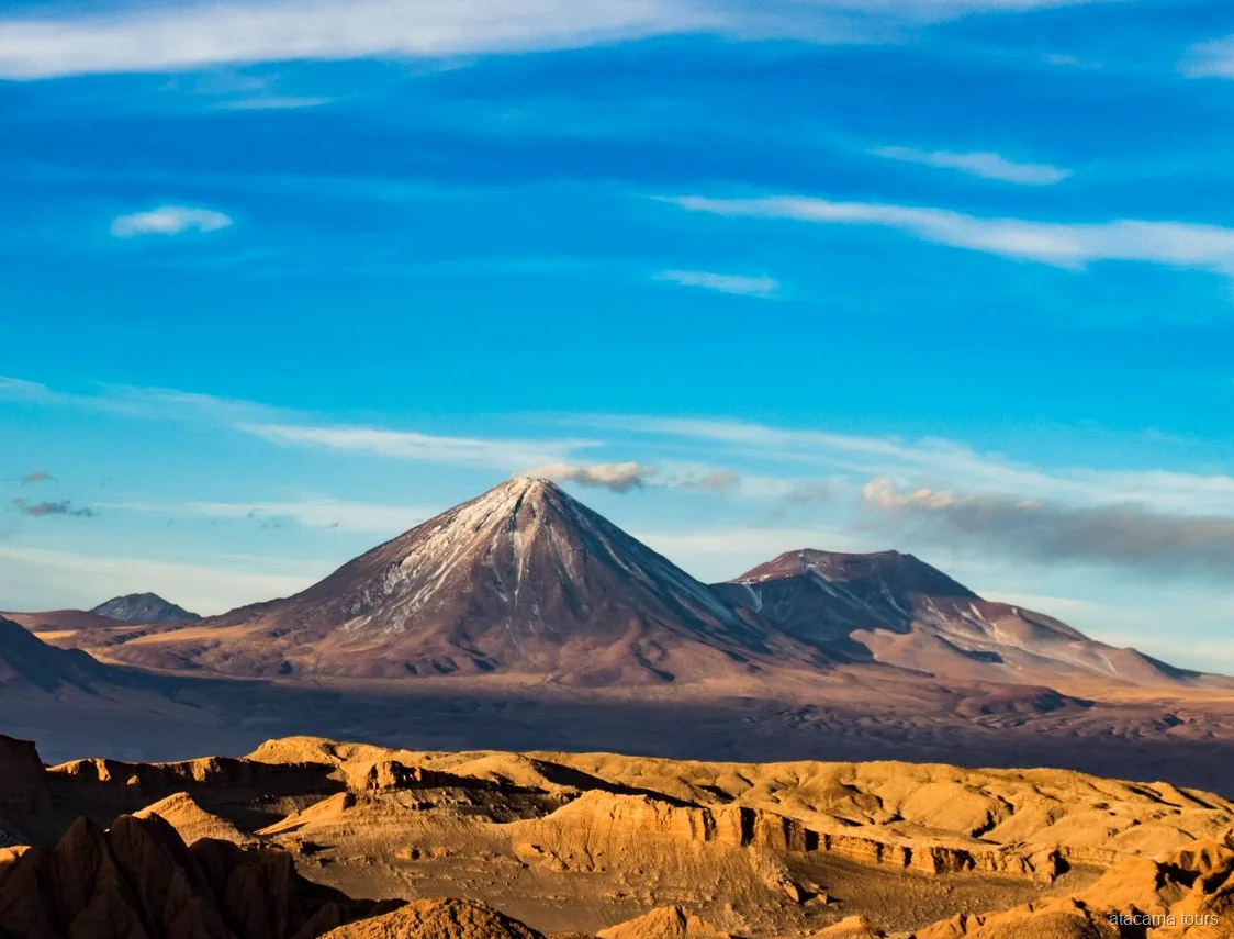 Atacama tours scenery, snow-streaked volcano cone towering over golden eroded desert ridges