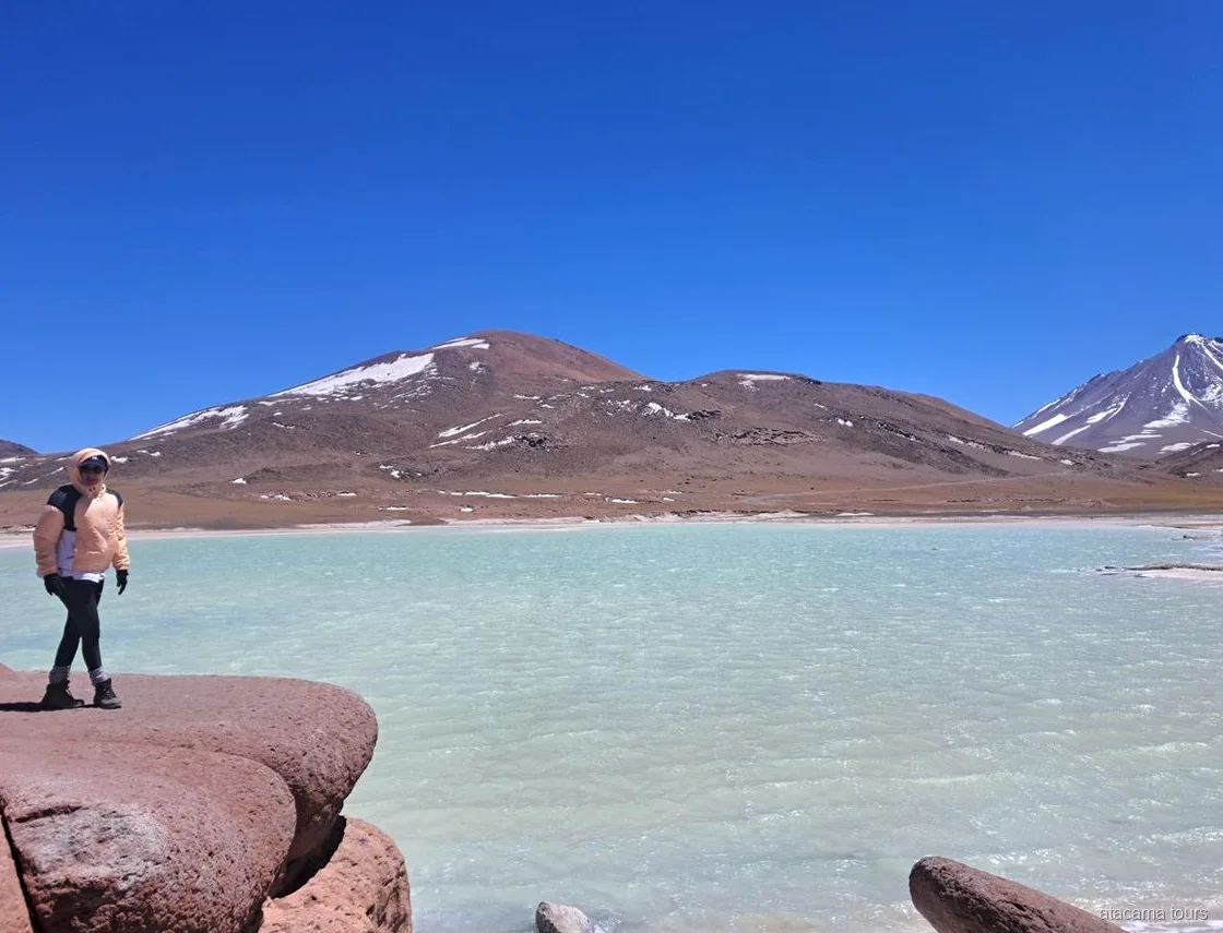 Atacama tours at Piedras Rojas, visitor standing on red rocks beside a turquoise lagoon and snowy peaks