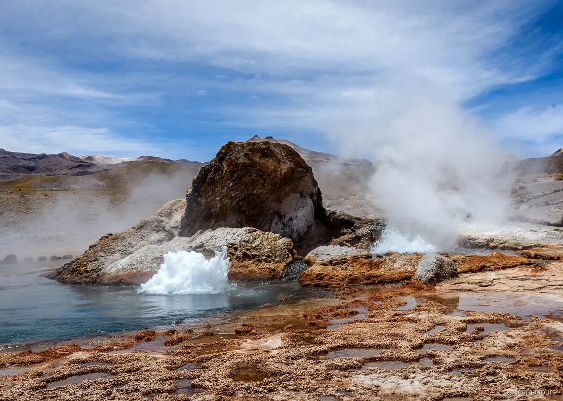 Tours en Atacama al Tatio, géiser burbujeando junto a una gran roca con vapor y terrazas minerales