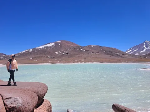 Tours en Atacama a Piedras Rojas, visitante sobre rocas rojas junto a una laguna turquesa y cerros nevados