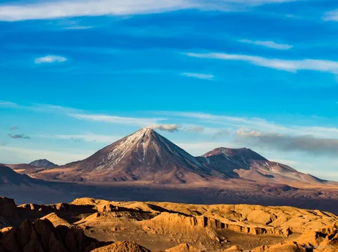 Paesaggio dei tour nel deserto di Atacama, cono vulcanico innevato sopra creste desertiche dorate ed erose