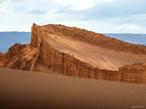Tour nel deserto di Atacama al Valle de la Luna, parete rocciosa inclinata a strati sopra una duna di sabbia