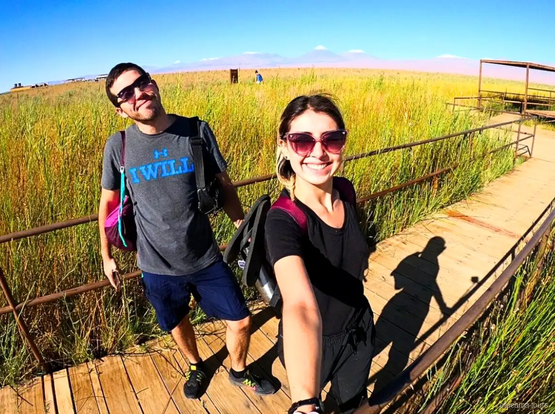 Pareja en tours en Atacama se hace un selfie en una pasarela de madera entre pastizales, volcanes al fondo